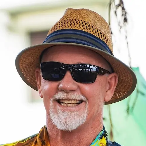 Greg Simpson smiling outdoors in a straw hat, dark sunglasses, and a colourful shirt, with greenery softly blurred in the background.