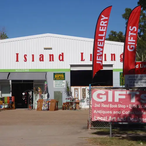 Storefront of Island Living, a white building with green trim, red flags reading “Jewellery” and “Gifts,” and various items displayed outside including furniture and décor.