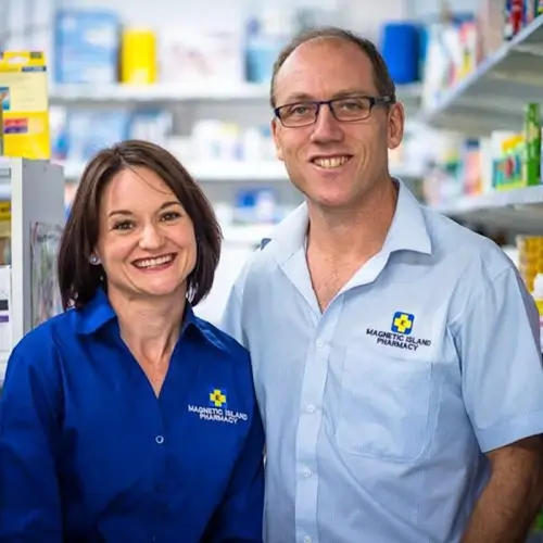 Sonia and Alan, owners of Magnetic Island Pharmacy, standing together inside the pharmacy wearing branded uniforms, with shelves of medicines and health products behind them.