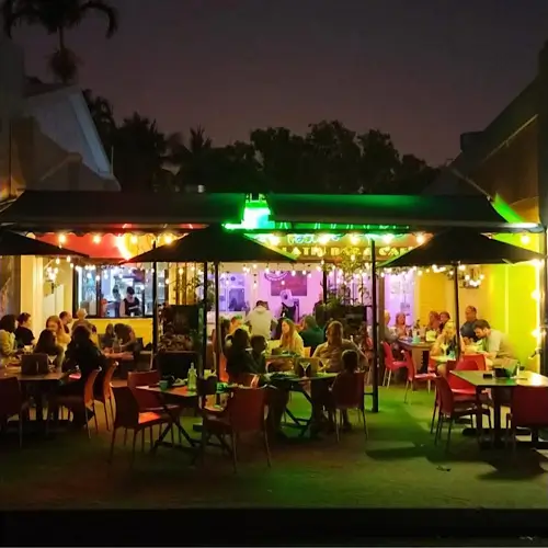 Noodies on the beach - an outdoor beachfront dining area at night with colourful lights, red chairs, and groups of people enjoying food and conversation.